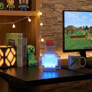 May include: A Minecraft-themed desk setup featuring a pixelated potion bottle lamp with blue and orange accents. A Minecraft mug, Creeper plush, and books are also visible. The computer screen displays a Minecraft game.