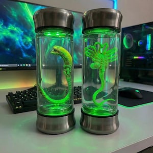 May include: Two glass bottles with stainless steel lids and bases, each containing a glowing green alien figure. The figures are detailed and appear to be submerged in a clear liquid. The bottles are illuminated with green light.
