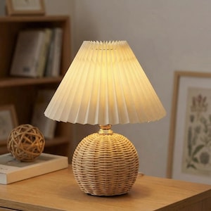 May include: A table lamp featuring a woven rattan base and a pleated, off-white lampshade. The lamp emits a warm light, suitable for a bedroom or living room. The spherical base and cone-shaped shade create a stylish design. The lamp is a decorative item.