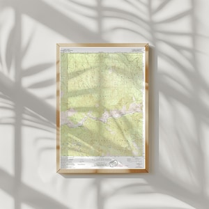 May include: A framed topographic map with a light wood frame. The map features shades of green and brown, depicting a mountainous region. The map is detailed with contour lines and elevation markings. The frame is rectangular and hangs on a white wall.