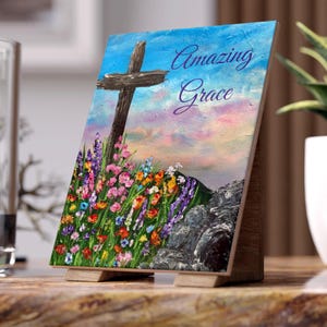 May include: A wooden easel displays a painting of a cross on a hill with colorful wildflowers. The sky transitions from blue to pink, with the words "Amazing Grace" in script. The painting is set on a wooden easel.