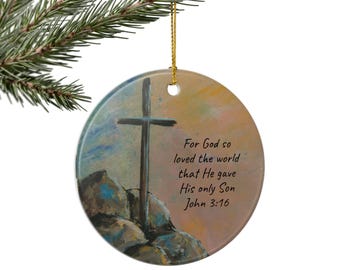 May include: Circular Christmas ornament featuring a painted cross on a rocky landscape, with the text "For God so loved the world that He gave His only Son John 3:16". The ornament is suspended by a gold string.