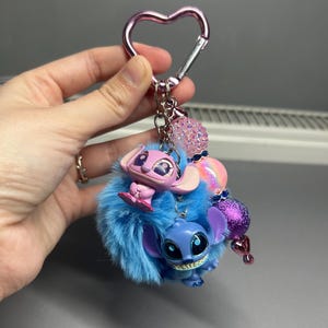 Peut inclure: Un porte-cl&eacute;s avec deux personnages de dessins anim&eacute;s, un bleu et un rose, attach&eacute;s &agrave; un pompon bleu duveteux. Le porte-cl&eacute;s a un fermoir en forme de c&oelig;ur rose et des perles d&eacute;coratives.