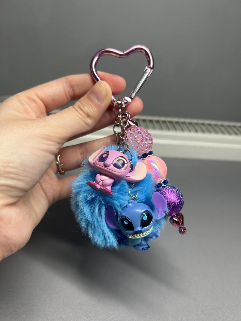 Peut inclure: Un porte-cl&eacute;s avec deux personnages de dessins anim&eacute;s, un bleu et un rose, attach&eacute;s &agrave; un pompon bleu duveteux. Le porte-cl&eacute;s a un fermoir en forme de c&oelig;ur rose et des perles d&eacute;coratives.