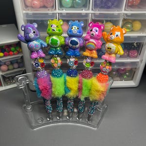 Könnte beinhalten: Eine Sammlung farbenfroher Stifte mit Care Bears-Figuren. Jeder Stift hat einen andersfarbigen Care Bear oben, mit einer flauschigen, regenbogenfarbenen Basis und einem glitzernden Stiftschaft. Die Stifte werden in einem transparenten Kunststoffständer präsentiert.