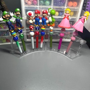 Op de afbeelding: Een verzameling pennen met figuren van populaire videogamekarakters. De pennen hebben kleurrijke kralenaccenten en karakterbeeldjes bovenop. De pennen worden gepresenteerd in een transparante plastic standaard.