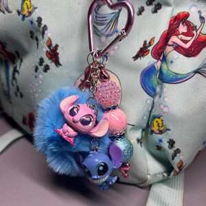 Peut inclure: Un porte-cl&eacute;s bleu et duveteux avec deux figurines de personnages de dessins anim&eacute;s, une rose et une bleue, attach&eacute;es &agrave; un mousqueton rose en forme de c&oelig;ur. Le porte-cl&eacute;s comporte &eacute;galement des perles roses et bleues. L'arri&egrave;re-plan est un sac &agrave; dos bleu clair avec un imprim&eacute; sir&egrave;ne.