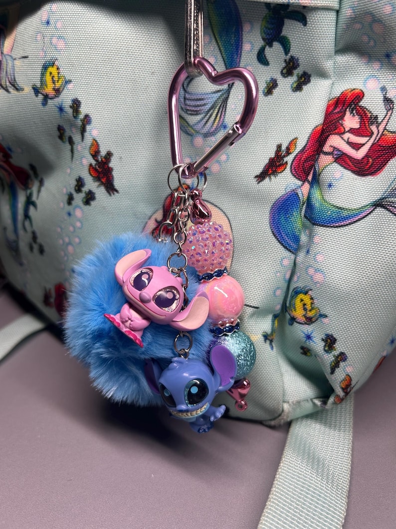 Peut inclure: Un porte-cl&eacute;s bleu et duveteux avec deux figurines de personnages de dessins anim&eacute;s, une rose et une bleue, attach&eacute;es &agrave; un mousqueton rose en forme de c&oelig;ur. Le porte-cl&eacute;s comporte &eacute;galement des perles roses et bleues. L'arri&egrave;re-plan est un sac &agrave; dos bleu clair avec un imprim&eacute; sir&egrave;ne.