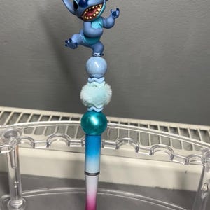 以下が含まれることがあります： 青いスティッチのフィギュアが上に付いた装飾的なペン。ペンの軸は青から白、ピンクへと変化し、青とターコイズのビーズが付いています。ペンは透明なプラスチックホルダーに展示されています。
