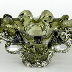 May include: An olive-green glass ashtray with a ruffled, organic shape. The ashtray has a textured surface and a series of rounded, curled edges. The glass has a slight transparency, allowing light to pass through.