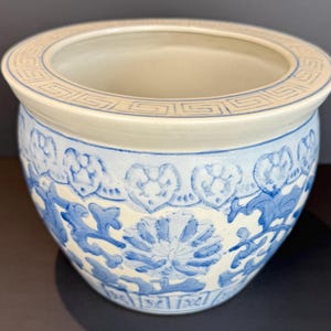 May include: A round ceramic planter with a cream interior and rim. The exterior features a blue and white floral design. The rim has a repeating geometric pattern. Suitable for indoor or outdoor use.