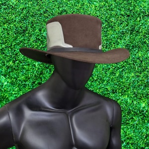 May include: A brown fedora-style hat with a wide brim and a black band. The hat features a light grey accent on the side. The hat is displayed on a black mannequin against a green background.