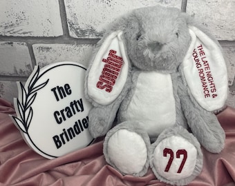 May include: A plush gray bunny with white accents on its ears and feet. The left ear features the red glitter text "Sombr" and the right ear reads "THE LATE NIGHTS & YOUNG ROMANCE". The left foot has the number "77" in red glitter embroidery.