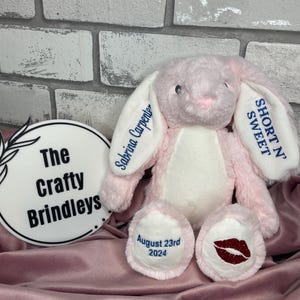 May include: A plush pink rabbit with white accents on its ears, chest, and feet. The left ear features the text "Sabrina Carpenter" and the right ear says "SHORT N' SWEET". The left foot displays "August 23rd 2024" and the right foot has a red lip print.