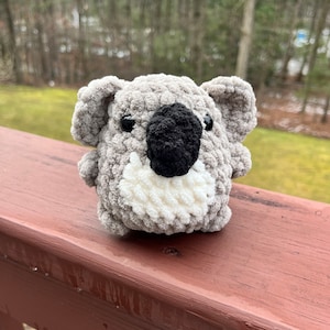 May include: A gray crocheted koala plush toy with black eyes and a black nose. The koala is sitting on a wooden surface.