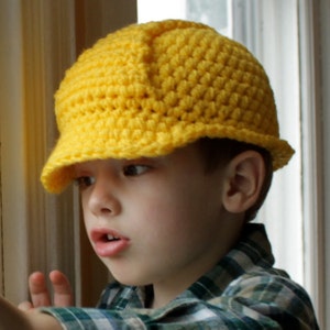 May include: A young child wearing a yellow crocheted construction worker hat. The hat has a brim and is made with a simple stitch pattern.