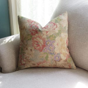 May include: A decorative throw pillow with a floral pattern in shades of pink, purple, and green on a cream background. The pillow is placed on a light-colored sofa, near a window.
