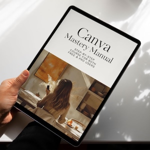 May include: A black tablet displaying the cover of the "Canva Mastery Manual." The cover features the title in a stylish font, with the subtitle "Step-by-Step Course for Both Free & Pro Users." The tablet is held by a person.