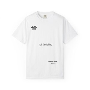 Könnte beinhalten: Weißes T-Shirt mit dem Text "PARIS 1980" über dem Satz "ngl. I'm falling" und den Worten "and I'm okay with it." in Schwarz. Das Shirt hat einen Rundhalsausschnitt und kurze Ärmel.