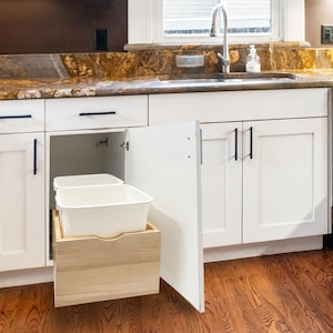 May include: White kitchen cabinet with an open door revealing a pull-out trash bin. The bin has two white plastic containers inside a wooden frame. The cabinet has black handles and is set against a granite countertop.