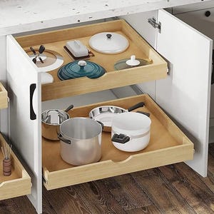 May include: Open white kitchen cabinet with two pull-out drawers. The top drawer holds cookware lids, a teal pot, and a butter dish. The bottom drawer contains stainless steel pots and pans, and a white pot.