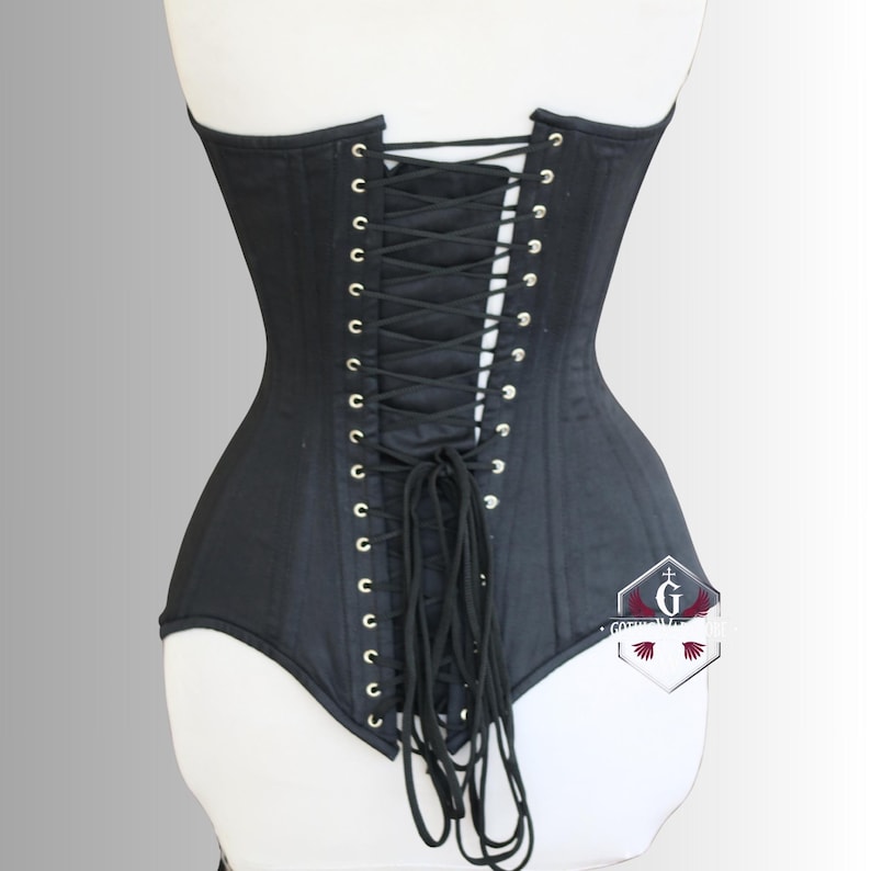 May include: A black corset with a lace-up back, featuring silver-toned eyelets. The corset is displayed on a white mannequin, showcasing its structured design and form-fitting silhouette. The garment has a smooth, matte finish.