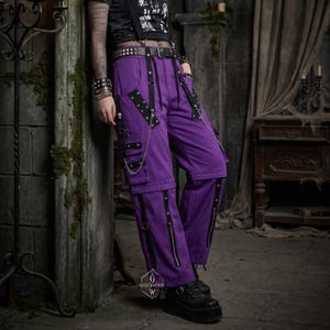 Può includere: Pantaloni cargo viola brillante con cinghie nere, catene in metallo argentato e occhielli metallici. I pantaloni includono tasche multiple, cerniere verticali sulla parte inferiore delle gambe e una cintura nera con fibbia argentata.