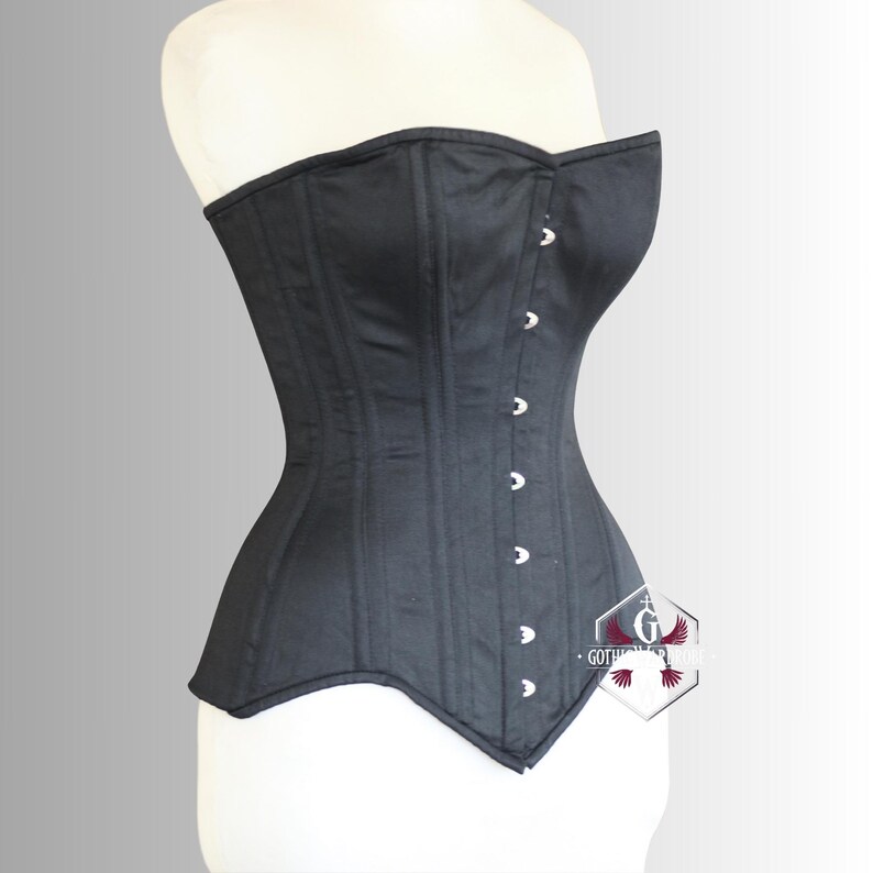 May include: A black corset with a structured, strapless design. The corset features vertical panels and is fastened with silver-colored clasps down the front. The garment is displayed on a white mannequin, showcasing its form-fitting silhouette.
