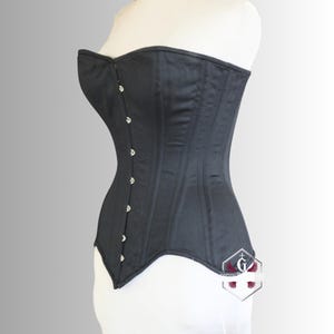 May include: A black corset with a structured silhouette. The garment features a series of silver-toned clasps down the front and vertical seams for shaping. The corset is displayed on a white mannequin, showcasing its form-fitting design.