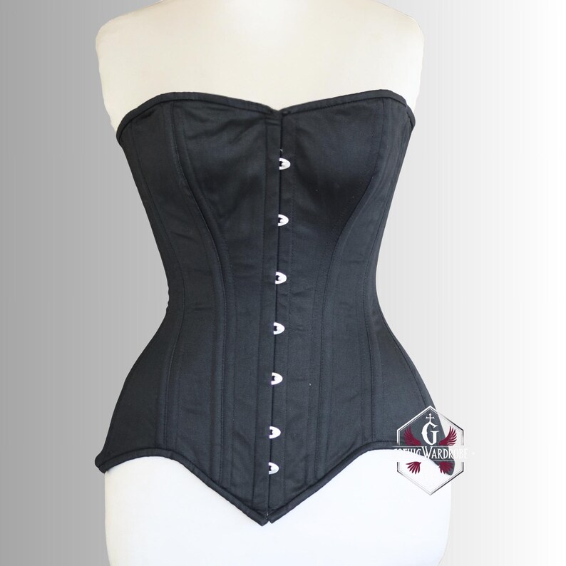May include: A black corset with a strapless design. The corset features a series of white clasps down the front and vertical seams for structure. The garment is displayed on a white mannequin.