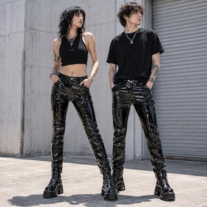 May include: Two individuals wearing black high-waisted pants with a glossy, patent leather-like finish. One individual wears a black sleeveless crop top and a silver necklace, while the other wears a black short-sleeved t-shirt and a silver chain.