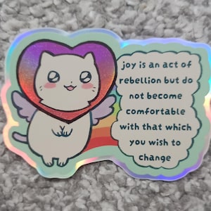 May include: A holographic sticker featuring a white cat with small wings inside a rainbow heart. The sticker includes the text: joy is an act of rebellion but do not become comfortable with that which you wish to change, set against a light green background.