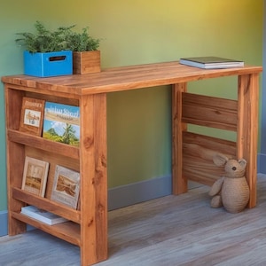 May include: A wooden desk with a natural finish, featuring a built-in bookshelf on one side. The desk has a rectangular top and a lower shelf for storage. A blue planter with greenery and a small stuffed animal are on the desk.