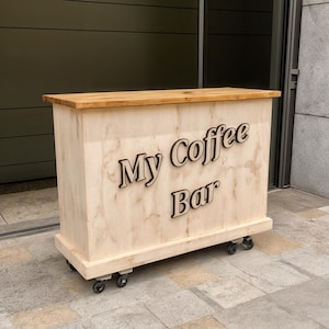 Op de afbeelding: Een lichtgekleurde houten koffiebar op wielen. De bar heeft een licht houten blad en de woorden "My Coffee Bar" staan op de voorkant. De bar is ontworpen voor mobiliteit en gemak.