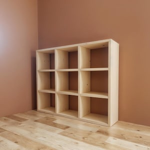 May include: A light-colored, nine-cube wooden shelf unit. The shelves are arranged in three rows of three, creating nine equal compartments. The unit is made of unfinished wood and sits against a brown wall and a wood-look floor.