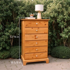 May include: A light brown wooden chest of drawers with six drawers and silver knobs. The chest is 53 1/4 inches tall, 24 3/4 inches wide, and 18 5/8 inches deep. A lamp with a white shade sits on top.