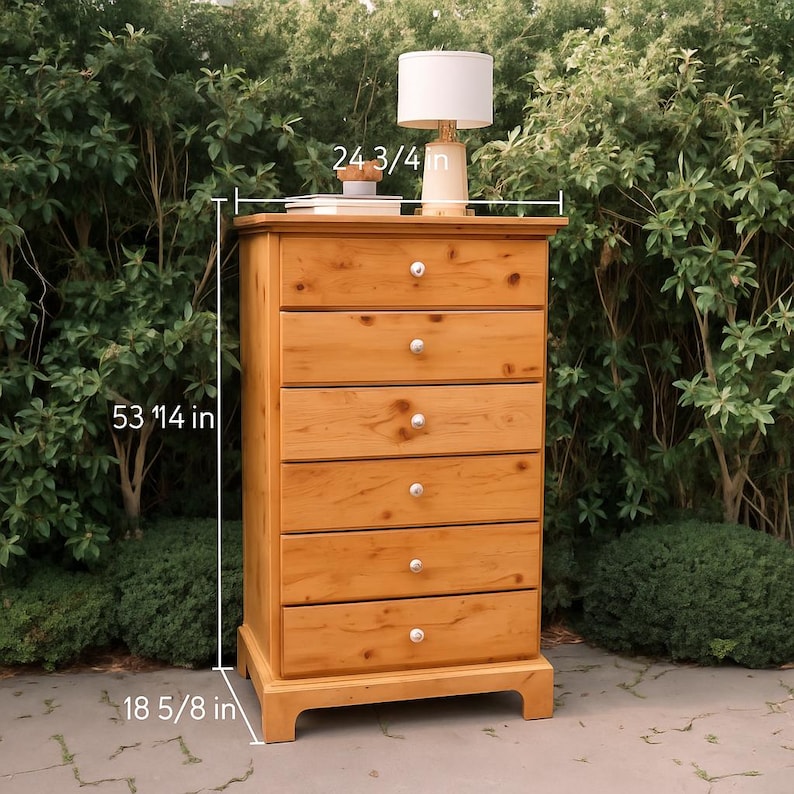 May include: A light brown wooden chest of drawers with six drawers and silver knobs. The chest is 53 1/4 inches tall, 24 3/4 inches wide, and 18 5/8 inches deep. A lamp with a white shade sits on top.