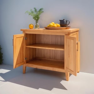 Könnte beinhalten: Ein helles Holzschrank mit zwei offenen Türen, die zwei Regale freigeben. Der Schrank hat eine flache Oberseite mit einem Tablett mit Zitronen, einer schwarzen Teekanne und einer Vase mit Grünzeug. Eine Orange befindet sich links von der Vase.