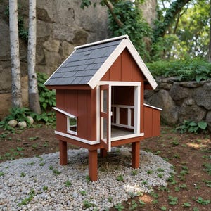 Könnte beinhalten: Ein kleiner Hühnerstall aus Holz, rotbraun lackiert mit weißen Zierleisten. Er hat ein graues Schindeldach, ein kleines Fenster und eine offene Tür. Der Stall steht auf vier Beinen und ruht auf einem Bett aus kleinen, grauen Steinen.