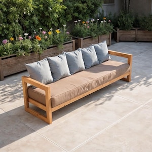 Könnte beinhalten: Ein hölzernes Outdoor-Sofa mit einem hellbraunen Kissen und fünf hellblauen Kissen. Das Sofa hat einen rechteckigen Rahmen und Armlehnen. Der Hintergrund zeigt Holzkästen mit bunten Blumen und Grün, was eine entspannende Umgebung schafft.