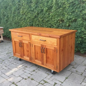 Puede incluir: Isla de cocina de madera con acabado natural. Cuenta con dos cajones y cuatro puertas de armario con tiradores negros. La isla está sobre ruedas y tiene una encimera de madera maciza.