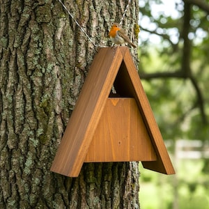 Könnte beinhalten: A-förmiges Vogelhaus aus Holz, das an einem Baum hängt, mit einem kleinen Vogel, der obenauf sitzt. Das Vogelhaus ist aus gebeiztem Holz gefertigt und hat eine dreieckige Form. Der Vogel hat eine rote Brust und braune Federn.