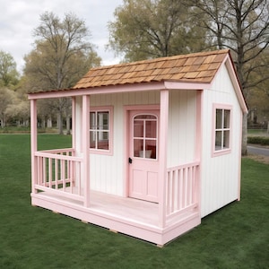 Könnte beinhalten: Ein weißes Spielhaus mit rosa Verzierungen, einer rosa Tür und einem braunen Schindeldach. Das Spielhaus hat eine kleine Veranda mit Geländer und steht auf einem grünen Rasen. Die Fenster sind rosa umrahmt.