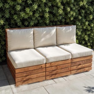 Puede incluir: Un sofá de exterior de tres plazas con estructura de madera y cojines color crema. El sofá está hecho de listones de madera y tiene un acabado de madera natural. Los cojines son de color crema claro. El sofá está en un patio con un seto verde al fondo.