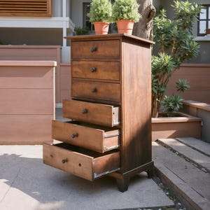 May include: A tall, wooden chest of drawers with five drawers, three of which are open. The chest is stained a warm brown color and has dark knobs. Two small potted plants sit on top. The chest is outdoors, with a tree and other plants in the background.