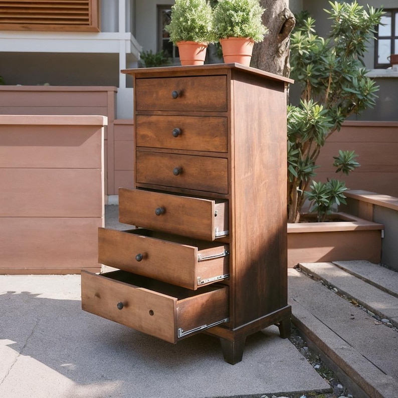 May include: A tall, wooden chest of drawers with five drawers, three of which are open. The chest is stained a warm brown color and has dark knobs. Two small potted plants sit on top. The chest is outdoors, with a tree and other plants in the background.