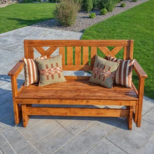 May include: A wooden outdoor bench with a natural finish, featuring a slatted seat and backrest with an X-shaped design. The bench is adorned with four decorative throw pillows in shades of brown, green, and cream, creating a cozy seating area.