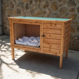 Puede incluir: Una casa de madera para mascotas con una parte delantera de malla metálica y una puerta de madera maciza. El techo es verde y el interior contiene una manta azul. La casa está elevada sobre cuatro patas y es de madera clara.