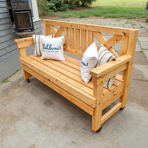 May include: A light-colored wooden bench with a slatted seat and back, featuring armrests and decorative cross bracing. Two white throw pillows with text and two striped pillows add comfort. The bench is on wheels for easy movement.