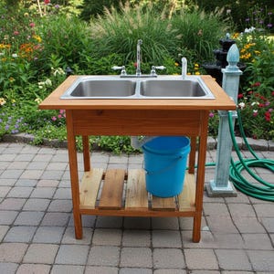Może przedstawiać: Drewniany zlew zewnętrzny z dwiema stalowymi umywalkami. Zlew ma drewnianą ramę i dolną półkę. Pod zlewem umieszczono niebieskie wiadro. W pobliżu zwinięty jest zielony wąż ogrodowy.